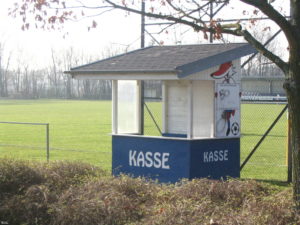 Ein blau-weißes Kassenhäuschen am Rande eines leeren Spielfeldes, auf dem das Wort KASSE und ein Fuchs mit Fußball gemalt ist. 