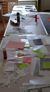 Auf einem ganz langen Tisch liegen wild durcheinander: Papierschnipsel, grüne und pinke Post-Its, Scheren und Kleber. 