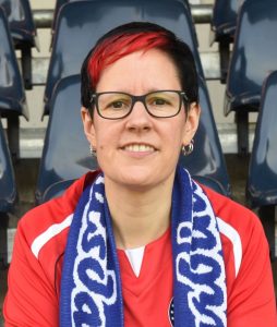 Carmen mit kurzen schwarzen Haaren, roter Strähne und Brille sitzt, bekleidet mit einem roten Turbinetrikot und Schal, im Stadion.