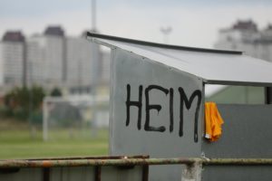 Ein Häuschen auf einem Sportplatz, auf dem der Schriftzug „HEIM“ gesprüht ist. Zudem hängt gelbes Trikot an dem Häuschen.