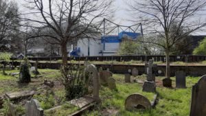Ein Friedhof im Vordergrund und im Hintergrund ragt das Stadion Stamford Bridge, die Heimspielstätte von Chelsea London, in den Himmel.