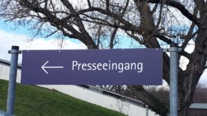 Ein blaues Schild zwischen zwei Metallpfosten zeigt einen weißen Pfeil mit dem Text „Presseeingang“.