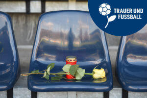Auf einem blauen Stadionsitz liegt eine gelbe Rose und ein rotes Grablicht steht auf dem Sitz. Oben steht in der Ecke in blauer Blase mit weißer Schrift "Trauer und Fußball".